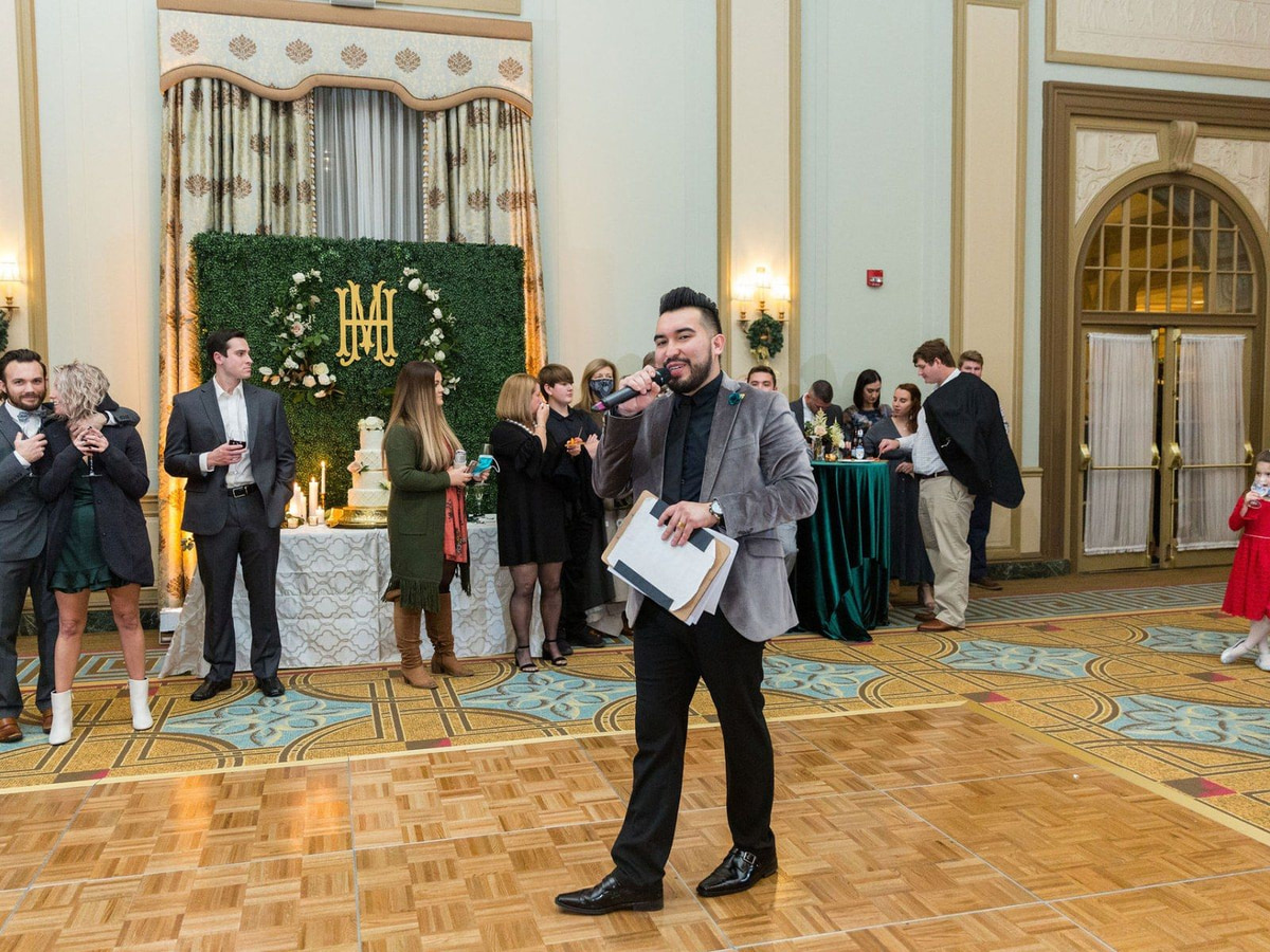 A SoundChoice DJ on the microphone on the ballroom dance floor, with reception guests and the wedding cake behind him