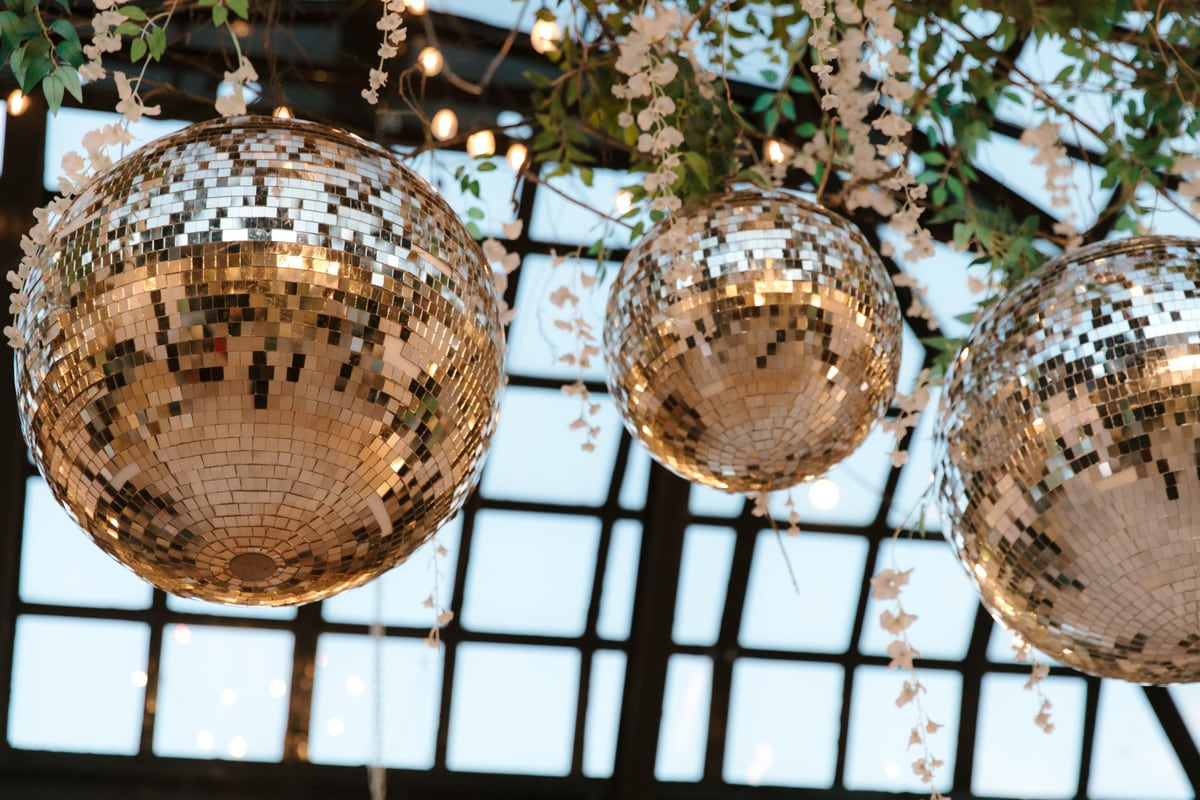 Three mirrored disco balls hanging beneath a pergola of fairy lights and greenery in a bright reception space